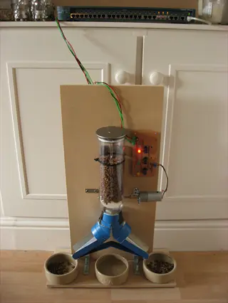 automatic cat feeder with kibble hopper, three bowls, and electronic components on a wooden board indoors