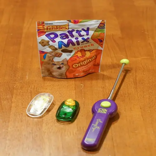 friskies party mix crunch cat treats bag with cartoon cat face, three clickers (clear, green, purple clikstik), demonstrating cat clicker training tools with treats
