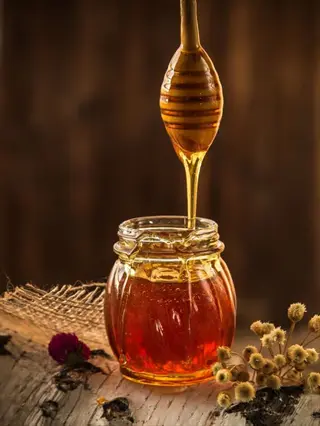 glass honey jar with golden honey dripping from a wooden dipper, surrounded by dried flowers and burlap on a rustic wooden surface