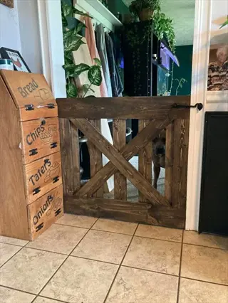 wooden dog safety gate blocking doorway with dog visible behind, next to storage unit labeled 'bread', 'chips', 'tatties', 'onions' in tiled room